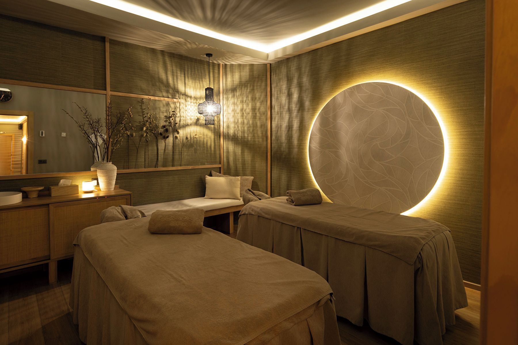 A spa room with two massage tables, a mirror, a sink, and a decorative plant.