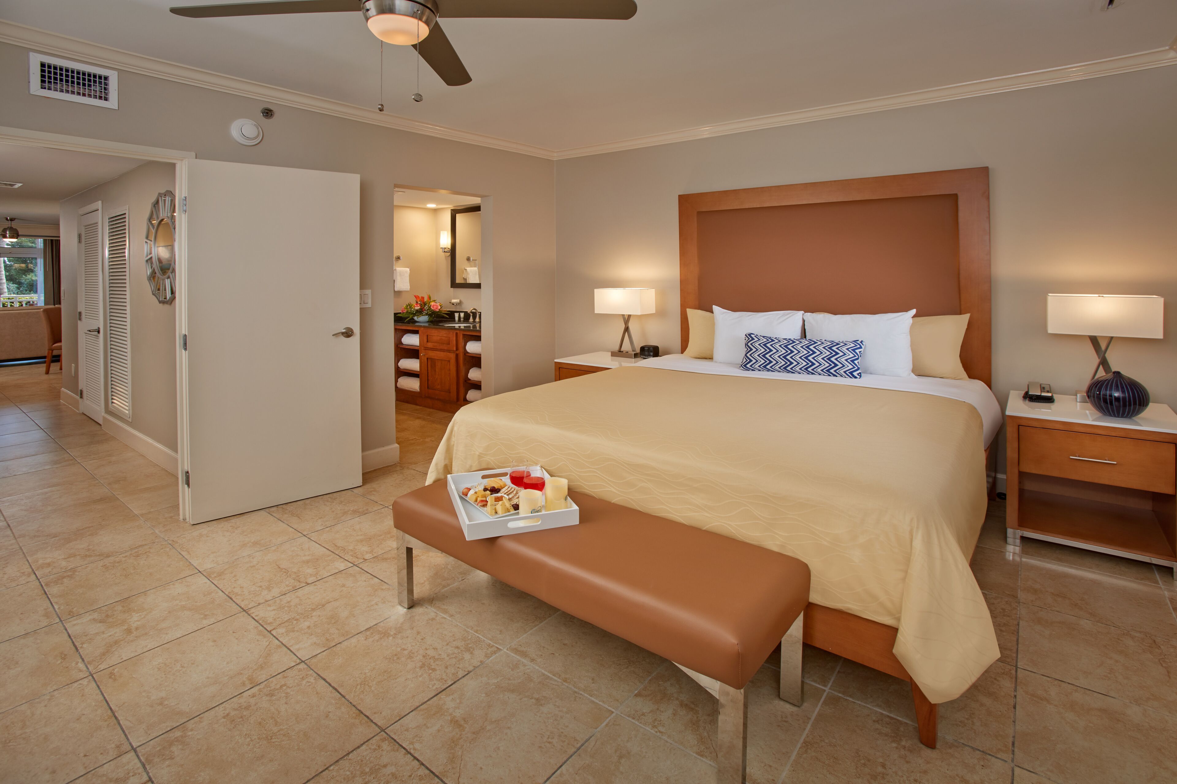Une chambre d’hôtel avec un grand lit, des tables de chevet, une banquette avec un petit-déjeuner, et une salle de bain visible à travers une porte ouverte.