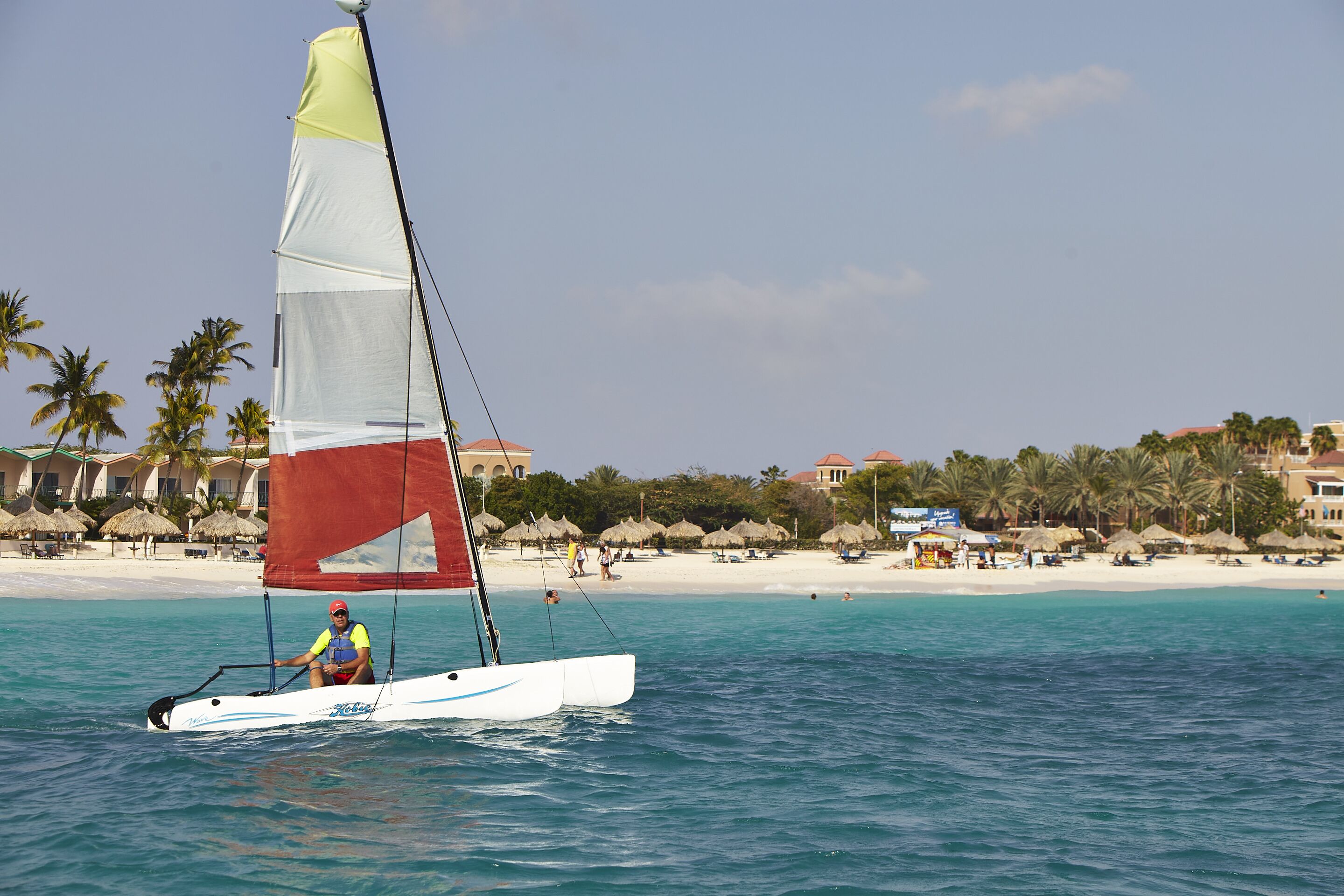 A sailboat on turquoise waters with a beach and palm trees in the background.