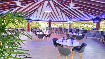 Zona de bar al aire libre con mesas y sillas redondas, una pantalla de televisión y un techo con vigas de madera y luces colgantes.