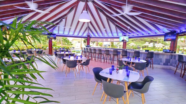 An outdoor bar area with round tables and chairs, a TV screen, and a ceiling with wooden beams and hanging lights.