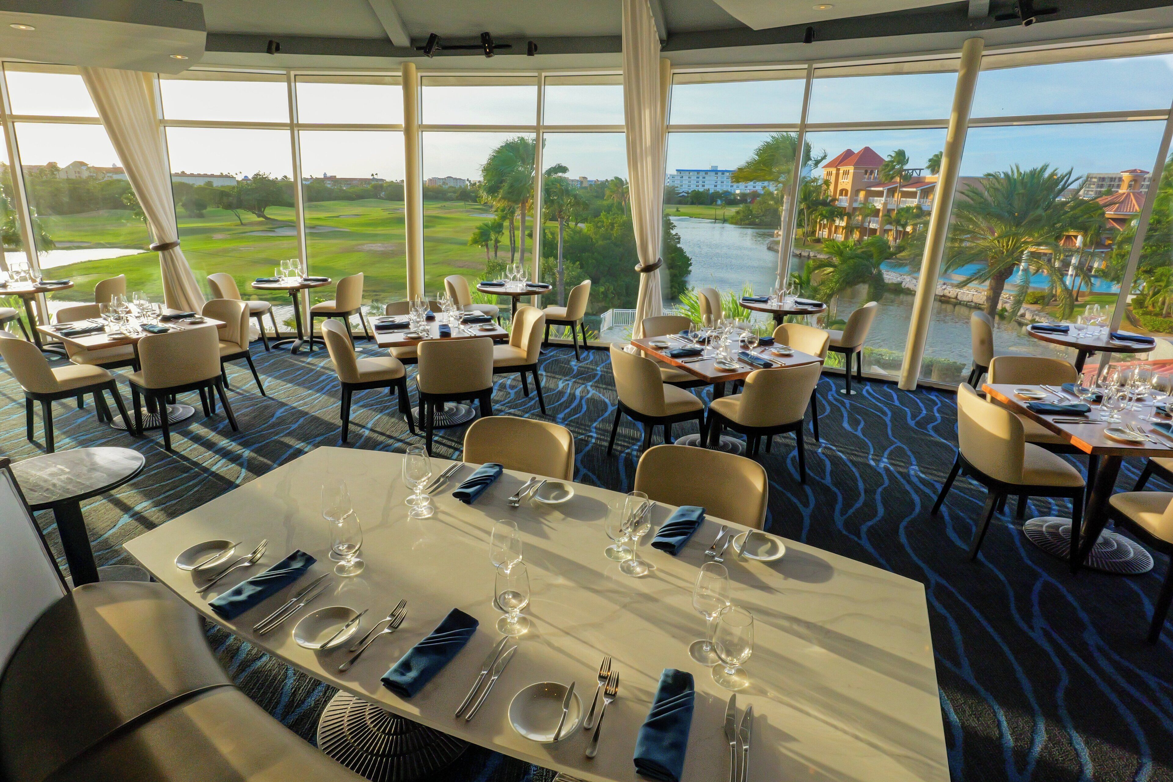 A restaurant with tables set for dining, large windows with a view of a golf course and water, and a carpeted floor.