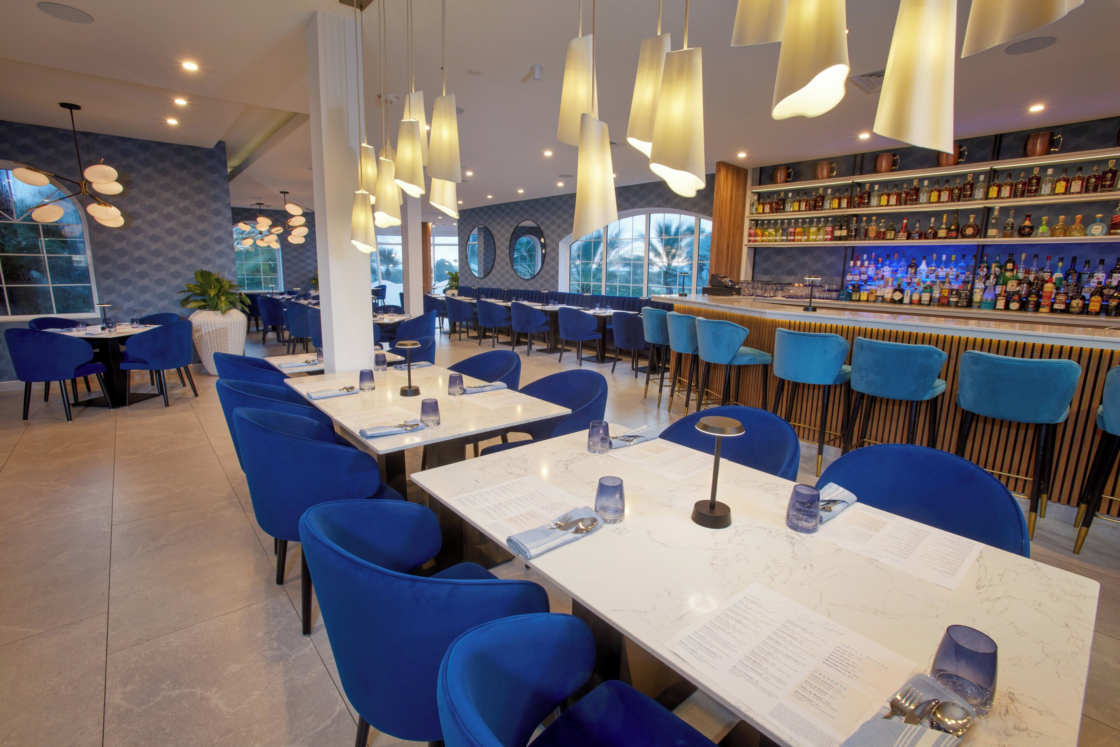 A restaurant with blue chairs, white tables, and a bar with shelves of bottles.