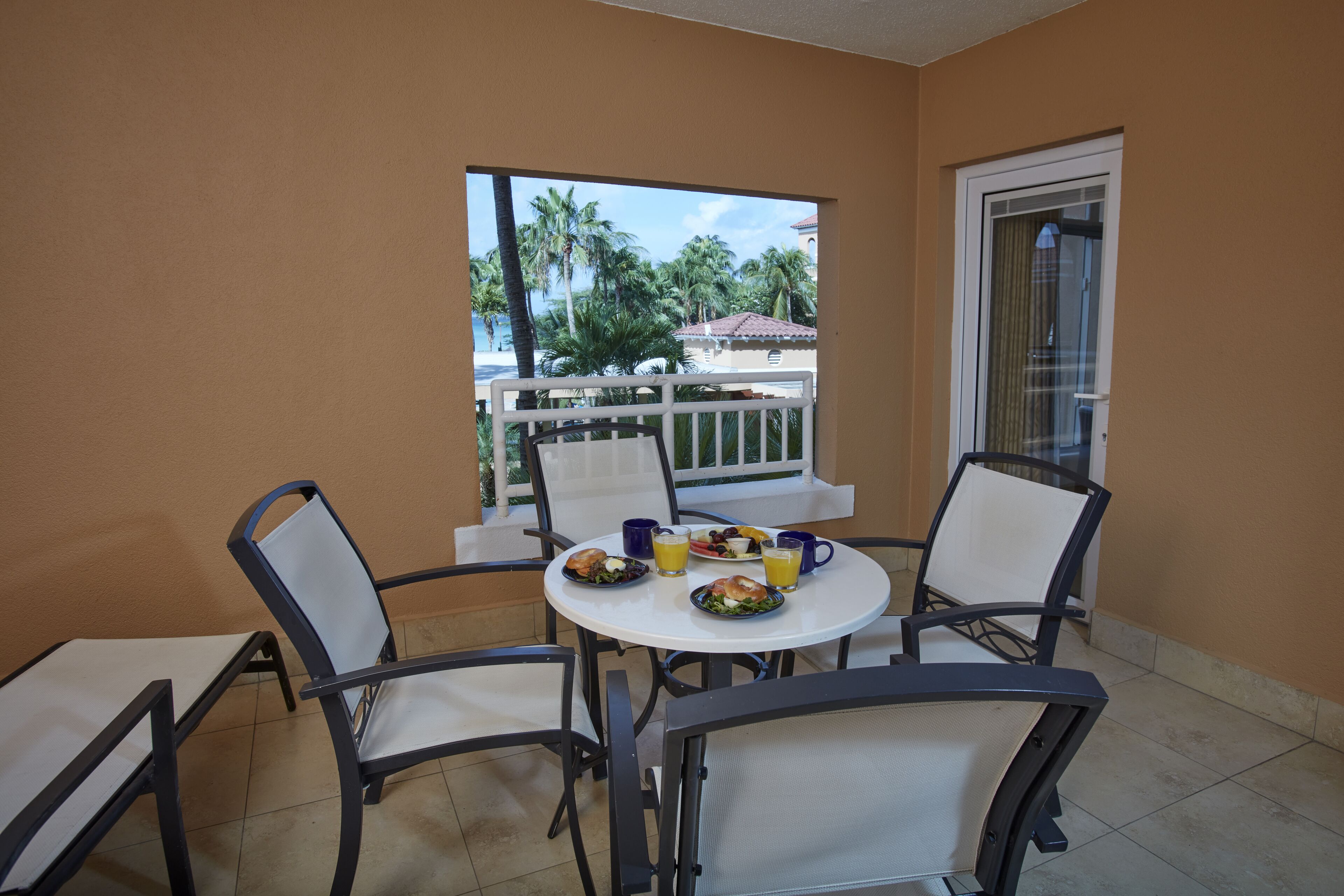 A balcony with a table set for two, chairs, and a view of palm trees and buildings.