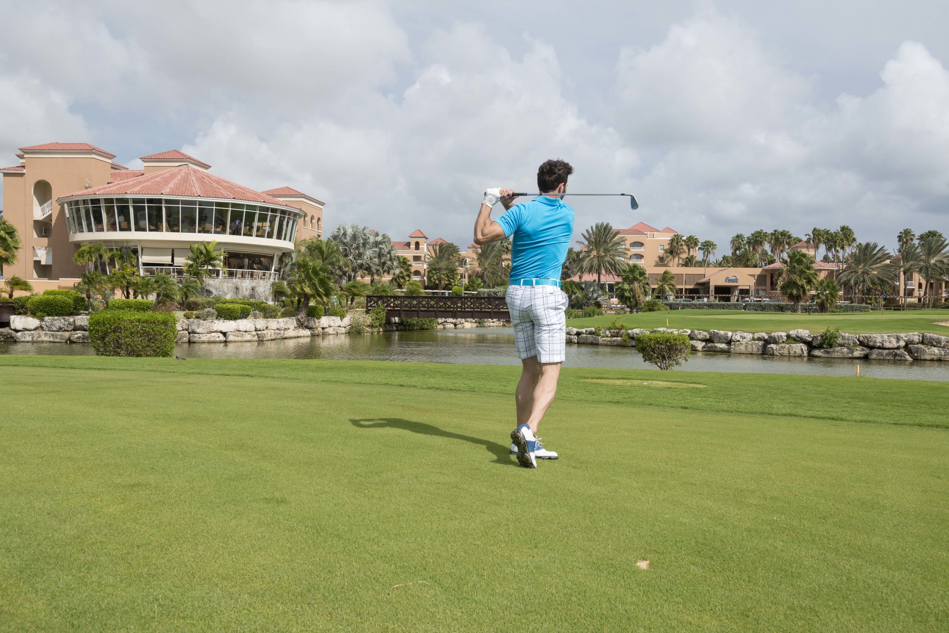 Un terrain de golf où l’on voit un homme portant un t-shirt bleu et un short à carreaux, prêt à frapper la balle avec son club de golf.
