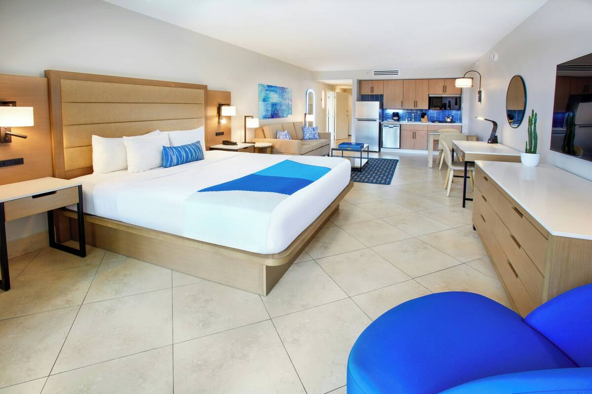 A hotel room with a large bed, a desk, a chair, a kitchenette, and a bathroom.