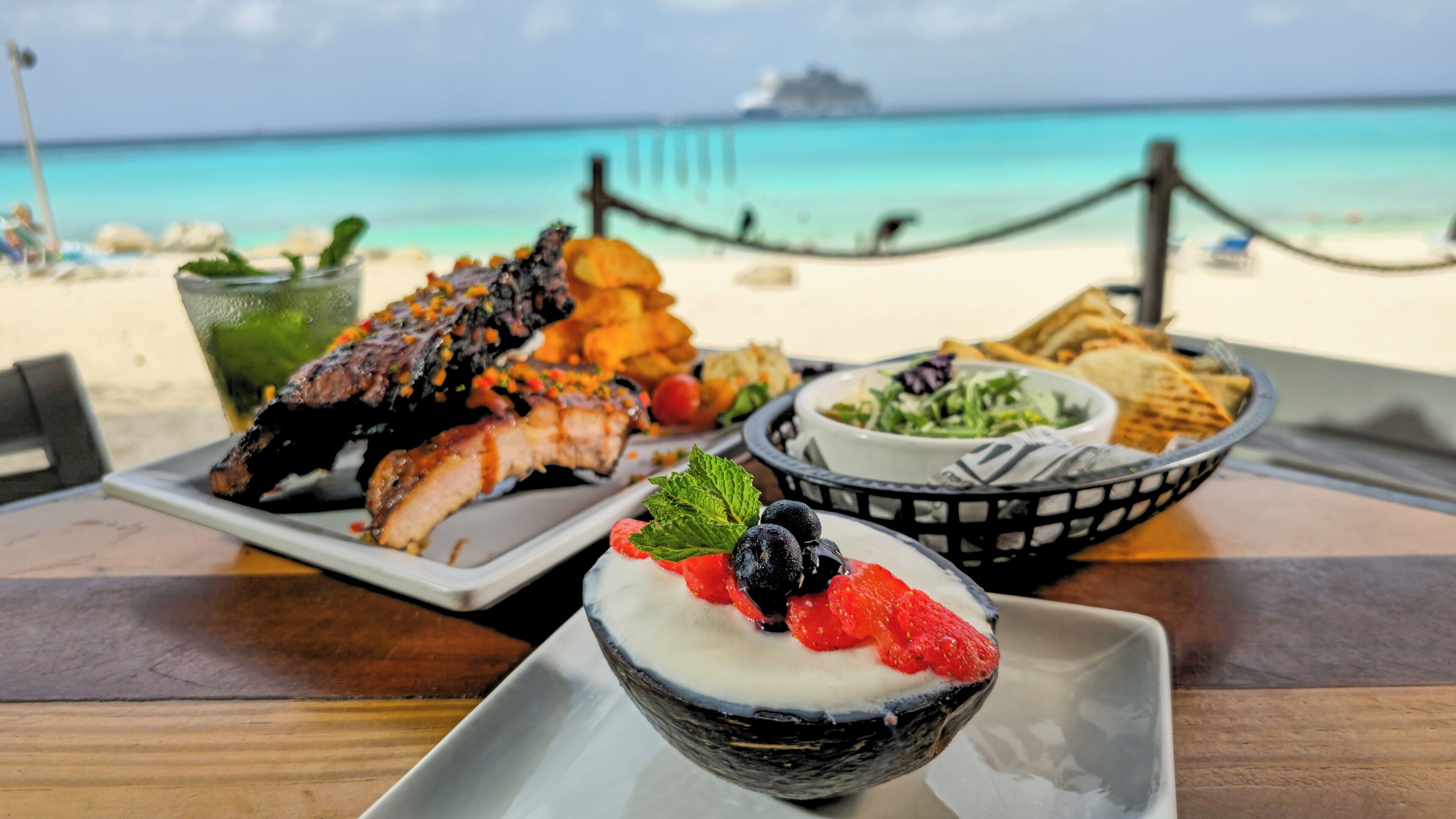 Ein Esstisch mit einer Auswahl an Meeresfrüchten, einer Salat-Schüssel und einer Nachspeise an einem Strand mit Blick aufs Meer.