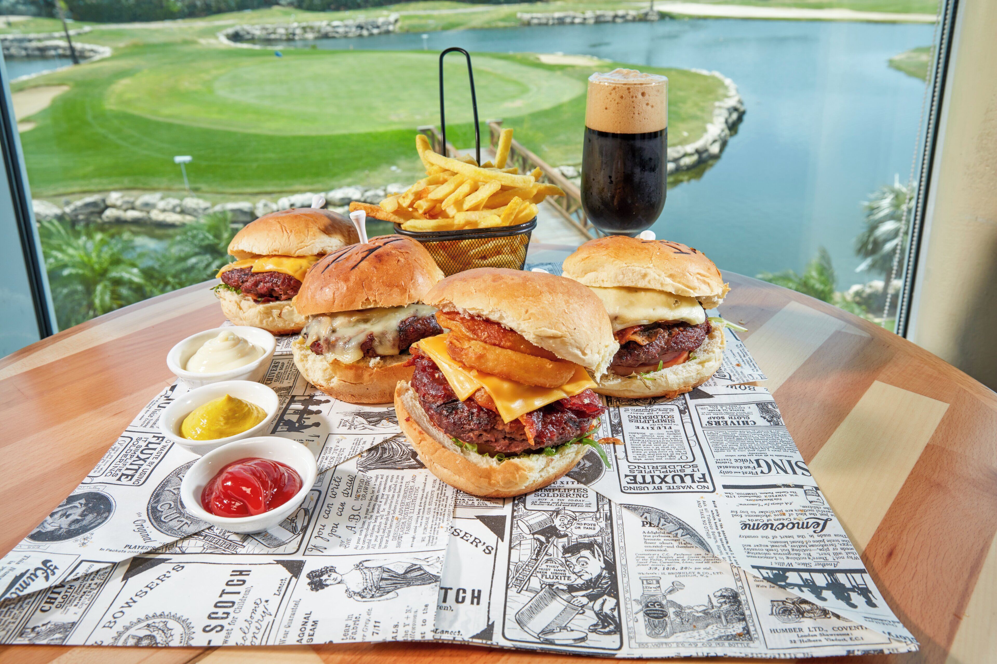 A table with burgers, fries, and drinks, with a golf course view in the background.