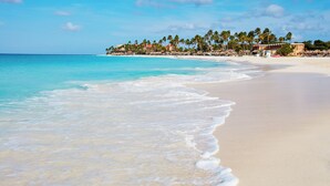 A beach with clear turquoise water, a sandy shore, and a resort area with buildings and palm trees in the background. - Divi Village Golf & Beach Resort (Oranjestad)
