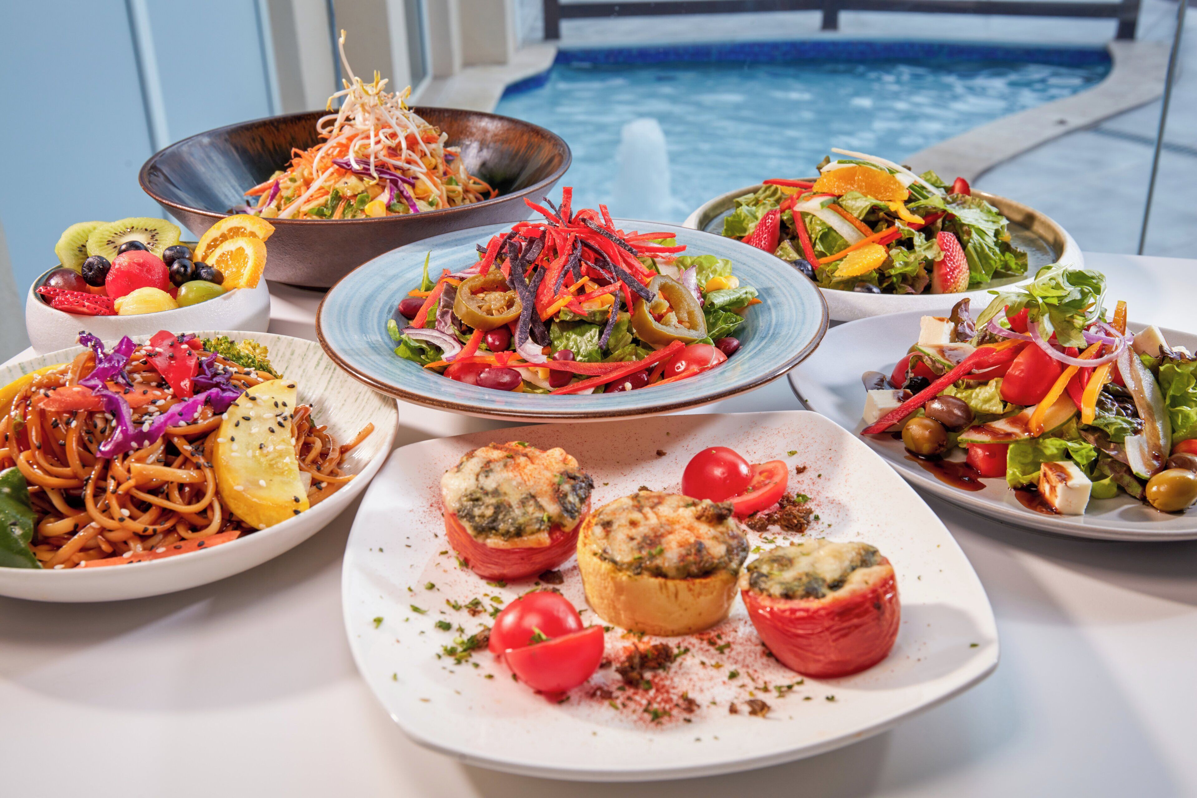 Variedad de platos en una mesa, incluyendo ensaladas y verduras rellenas, con una piscina al fondo.