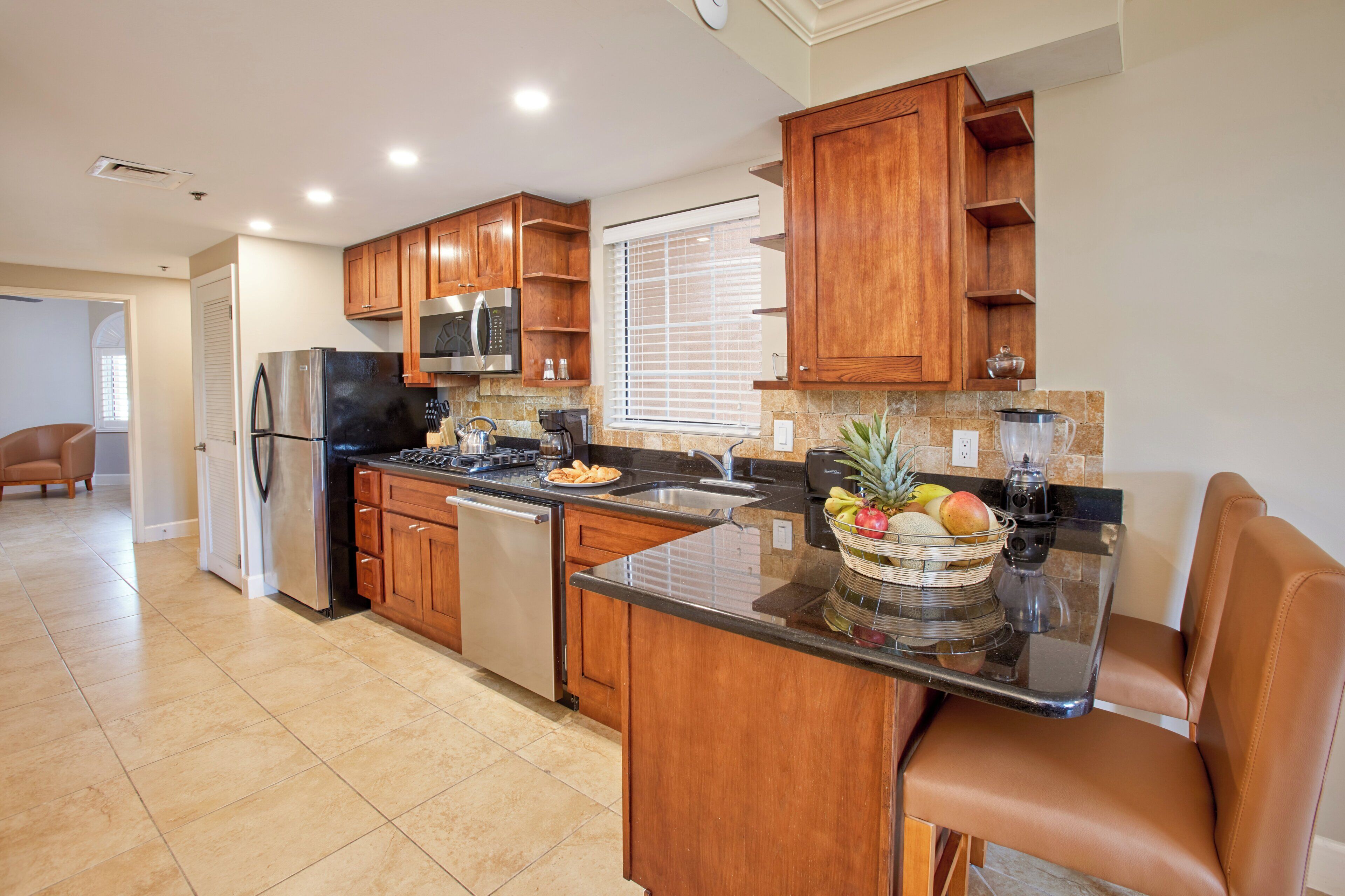 A modern kitchen with wooden cabinets, stainless steel appliances, a granite countertop, and a fruit basket.