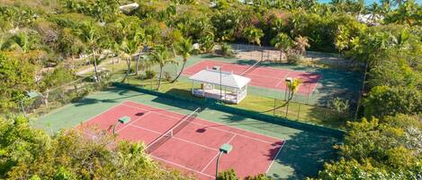 A tennis court complex with multiple courts, surrounded by lush greenery and a view of the ocean.