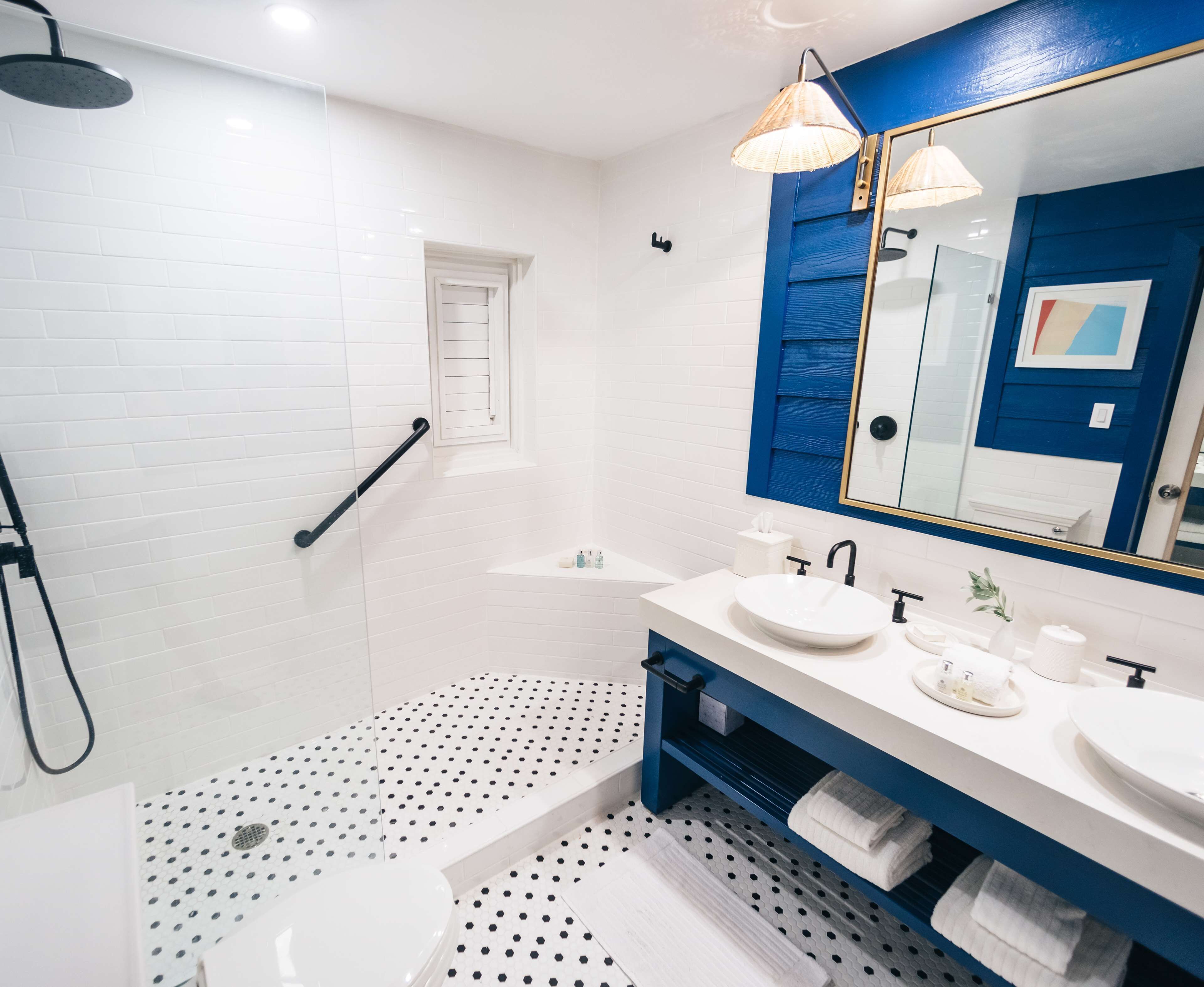 A modern bathroom with white tiled walls, a blue accent wall, a large mirror, two sinks, a shower, and a polka dot floor.