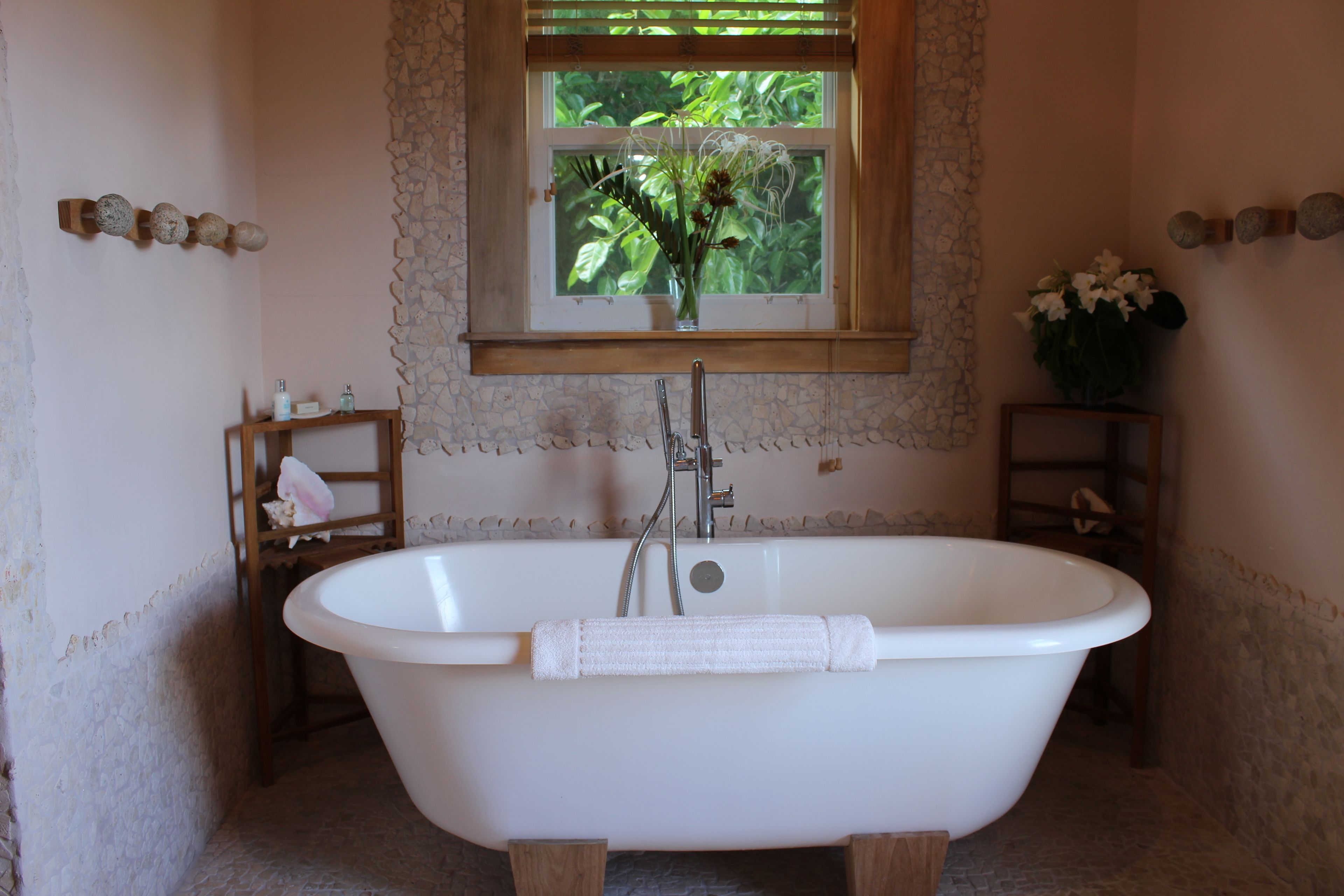 A bathroom with a freestanding bathtub, a window with a view of greenery, and a wooden shelf with toiletries.