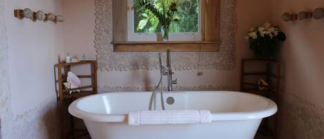 A bathroom with a freestanding bathtub, a window with a view of greenery, and a wooden shelf with toiletries.