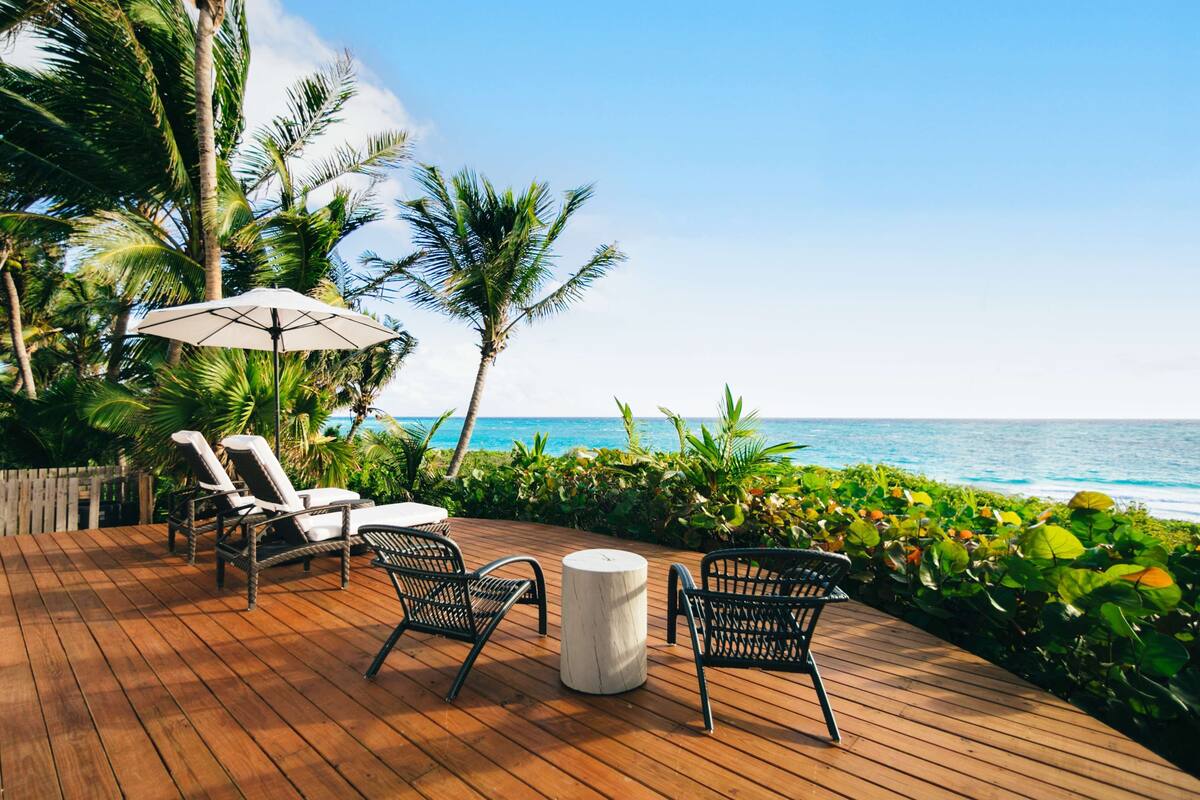 A wooden deck with lounge chairs and a table, overlooking a beach with palm trees and ocean views.