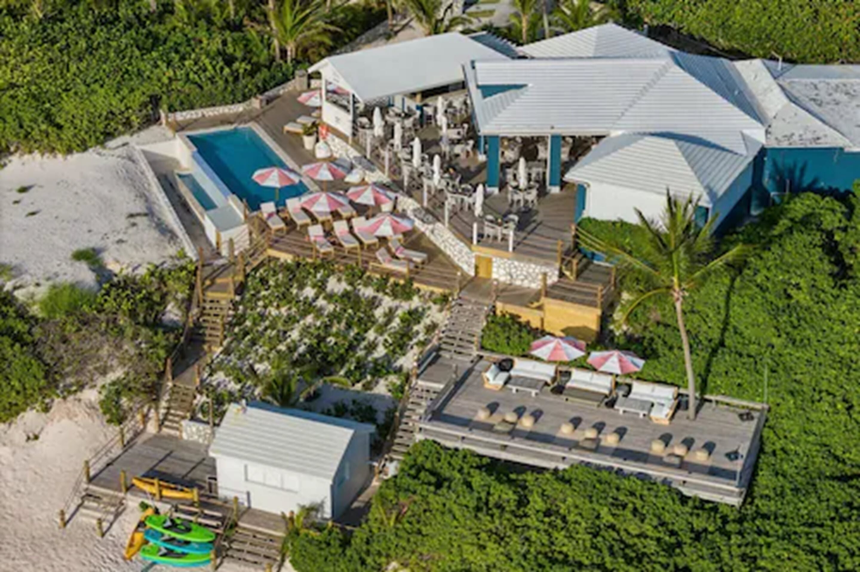 An aerial view of a resort with a swimming pool, beachside seating, and a covered area with umbrellas.
