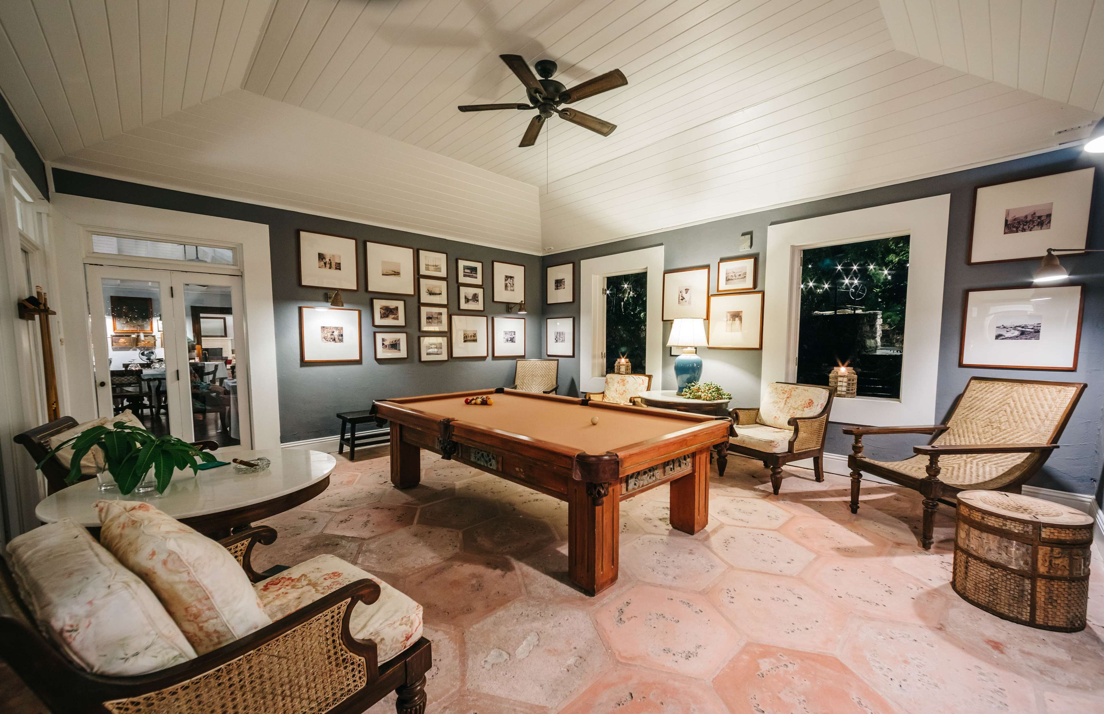 A pool table in a room with a ceiling fan, framed pictures on the walls, and wicker furniture.