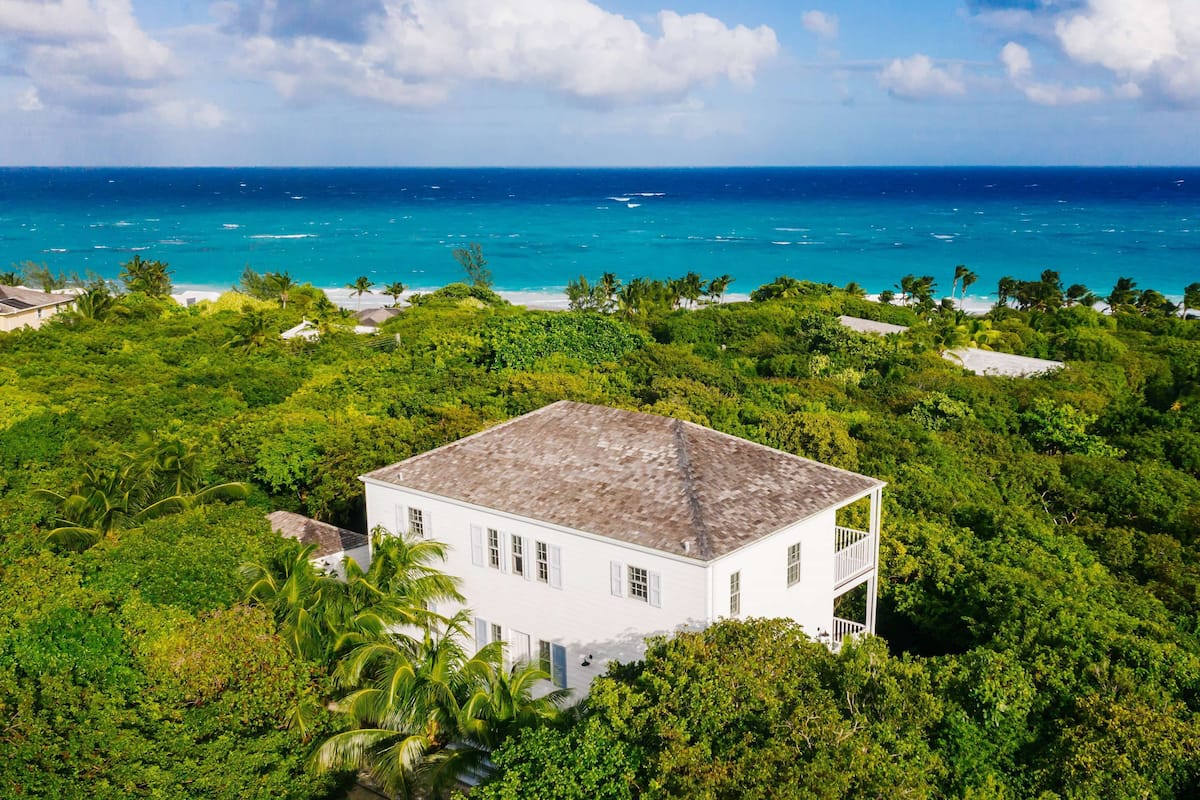 A white building surrounded by dense tropical vegetation, overlooking a turquoise ocean with visible boats.