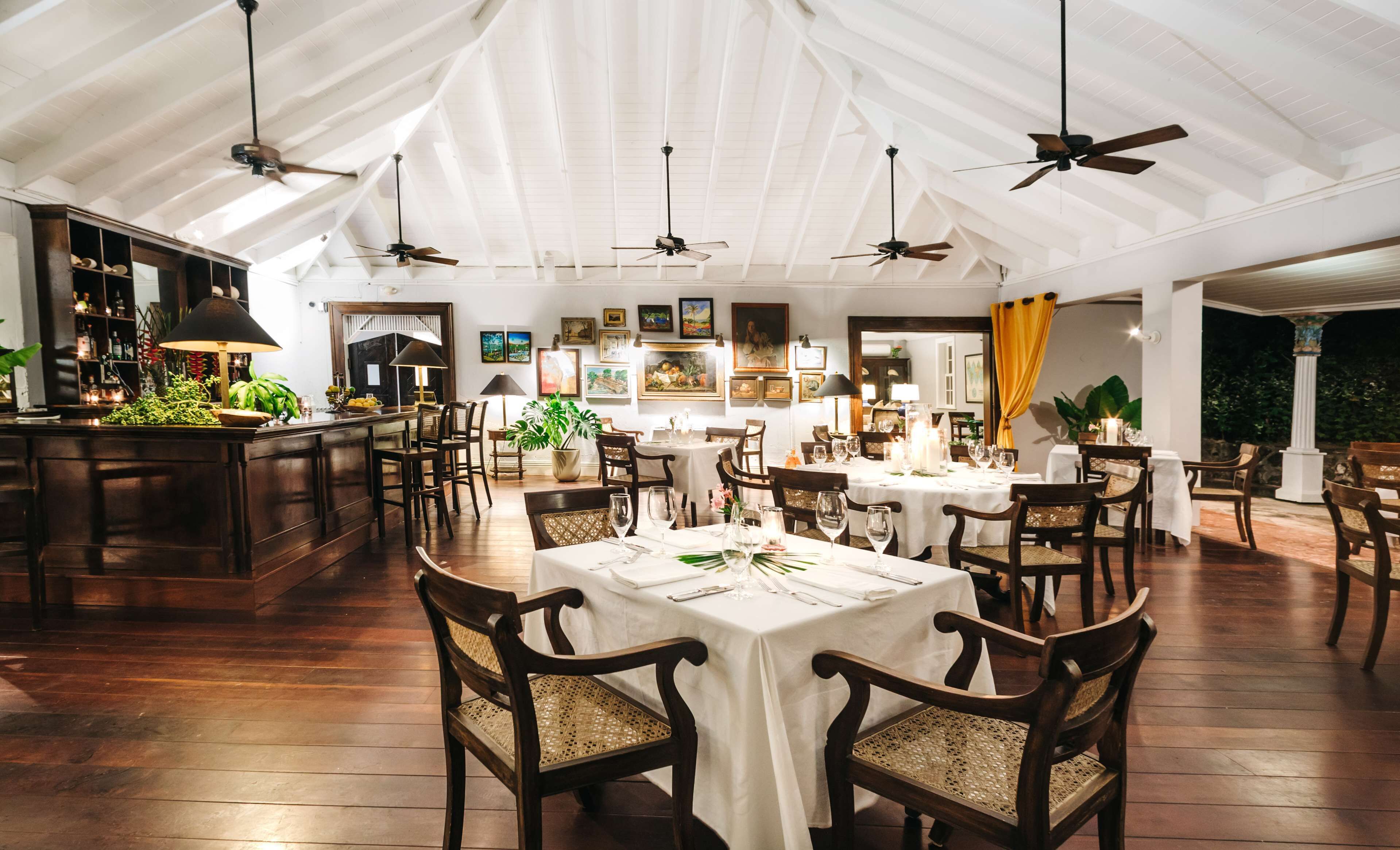 A spacious dining area with high ceilings and multiple ceiling fans, wooden tables and chairs, and a bar area with a counter and shelves.