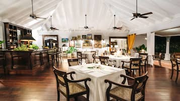 A spacious dining area with high ceilings and multiple ceiling fans, wooden tables and chairs, and a bar area with a counter and shelves.