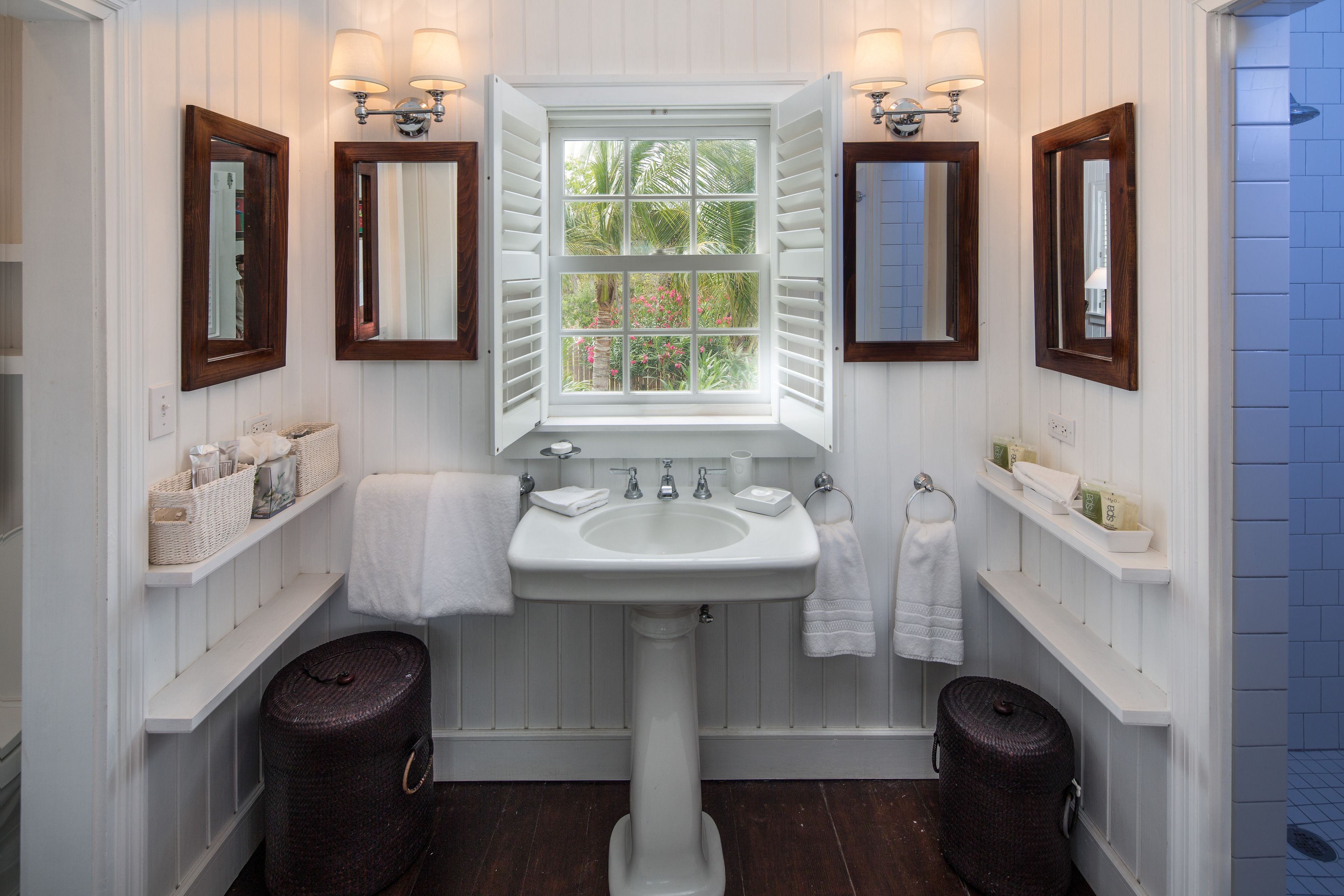 A bathroom with a white pedestal sink, two mirrors, a window with shutters, and wooden barrels.