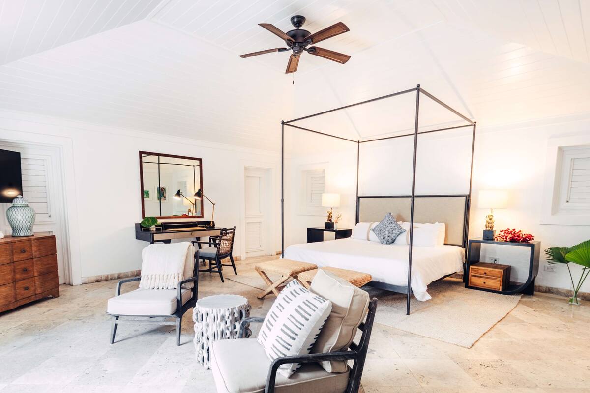 A spacious bedroom with a four-poster bed, a ceiling fan, a large mirror, and a seating area with chairs and a small table.