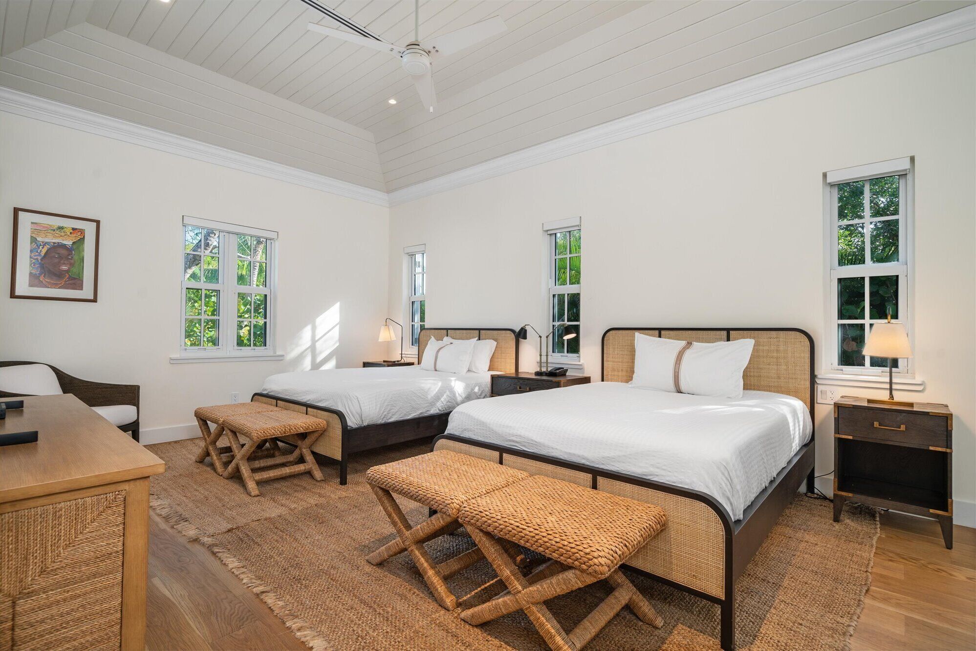 A bedroom with two beds, a desk, and a chair. There are windows with views of greenery.