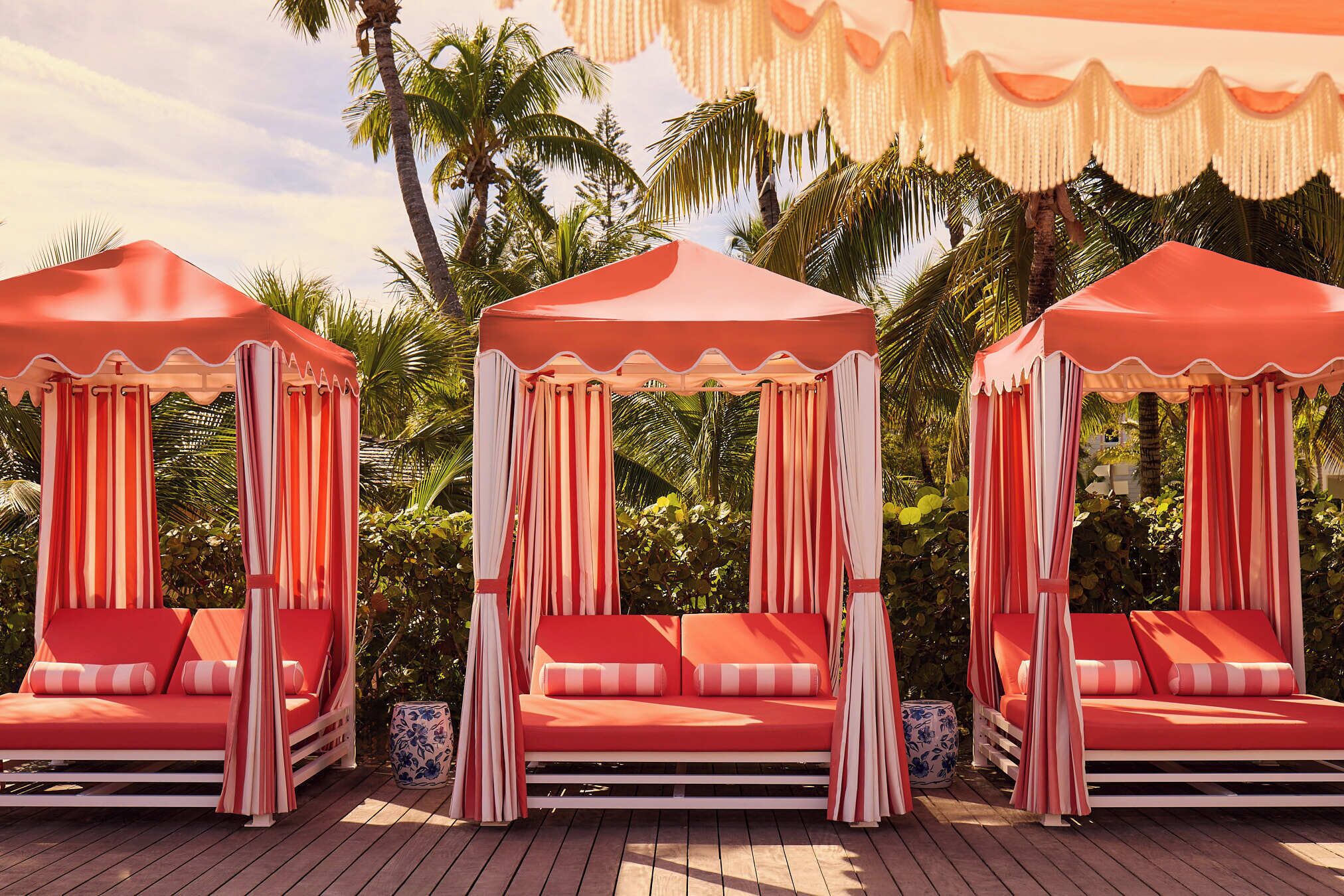 Three red canopy beds with white curtains, set on a wooden deck with palm trees in the background.