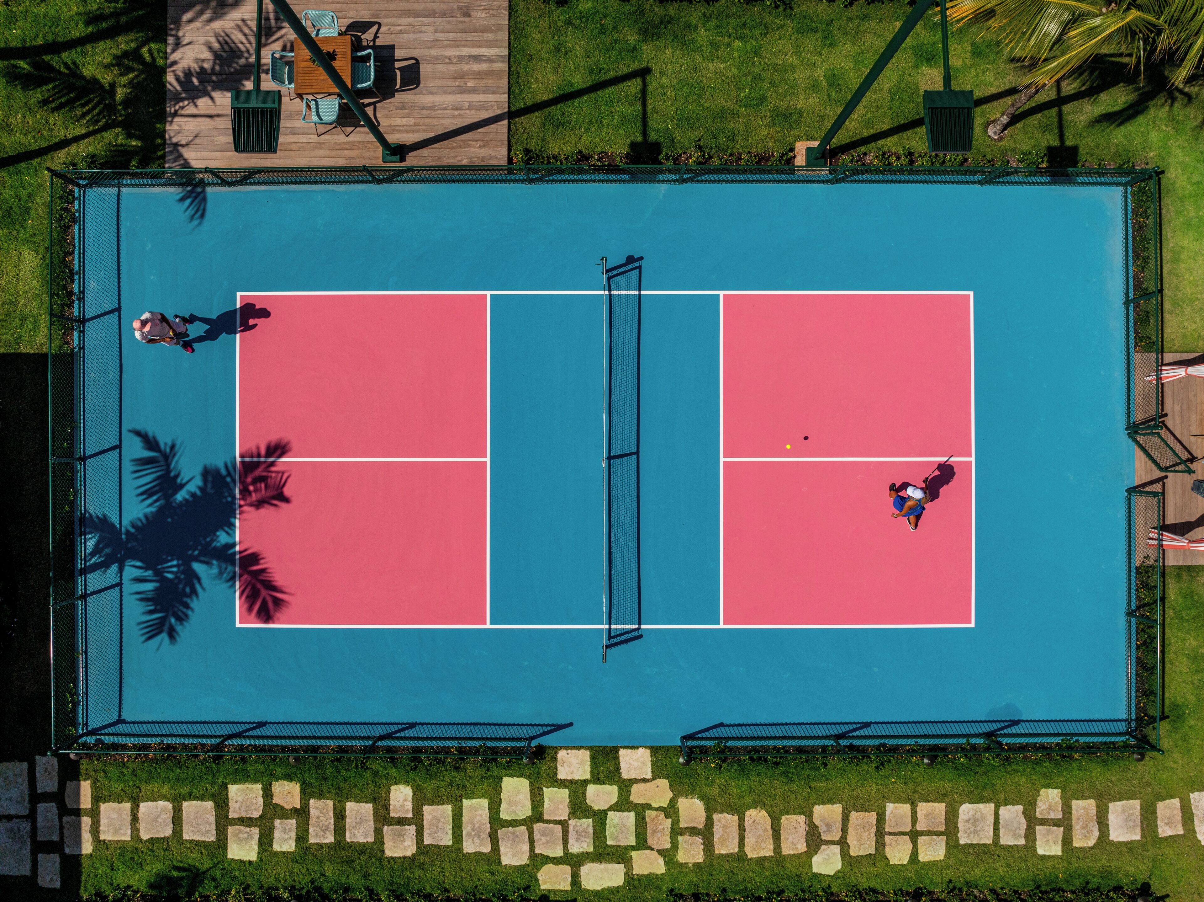 A tennis court with two players, surrounded by a fence and palm trees.