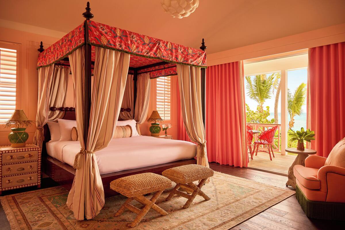 A four-poster canopy bed with a red and beige canopy, a wooden nightstand with a lamp, and a view of the outdoors through a sliding glass door.