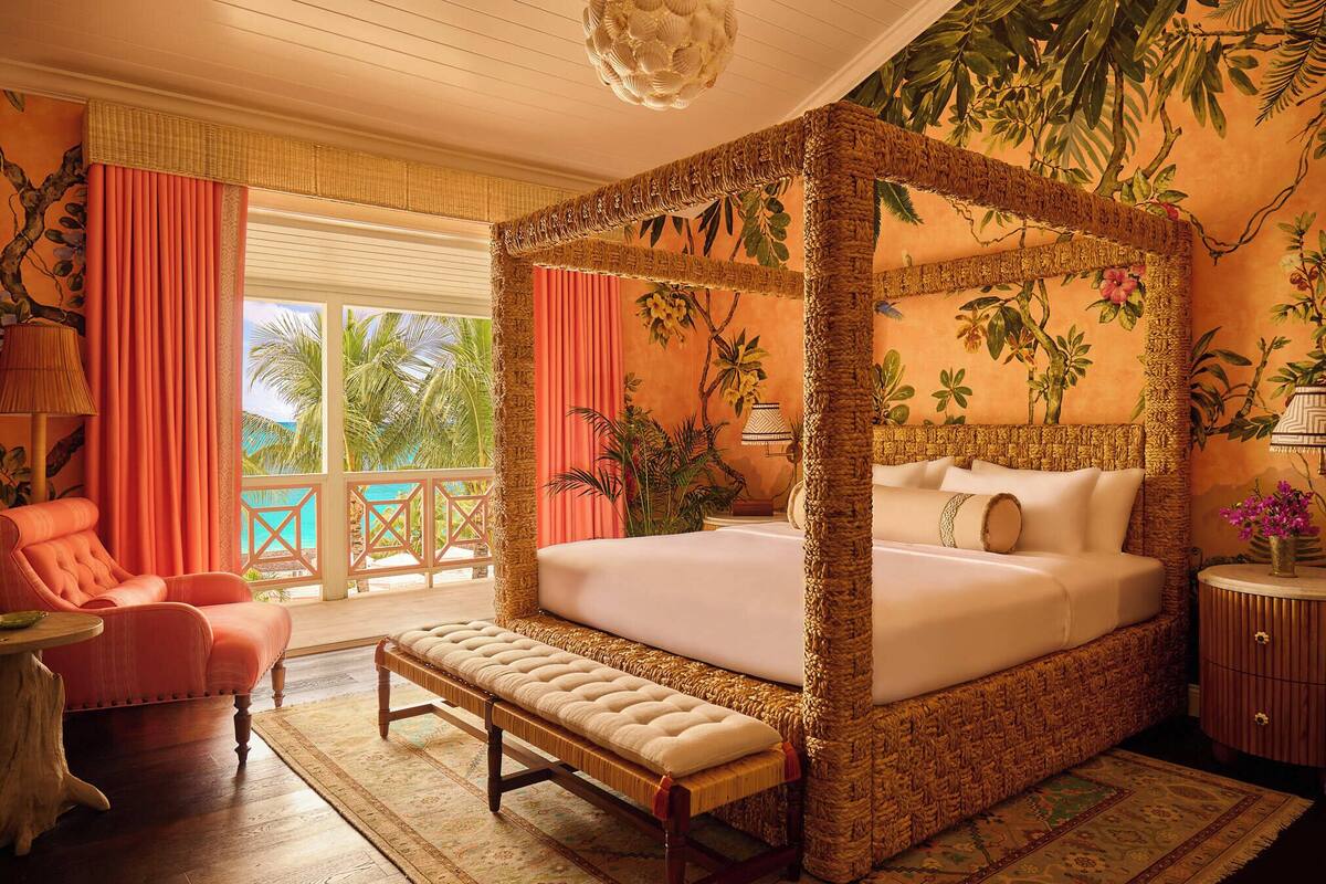 A four-poster bed with a woven frame, a bench, a pink armchair, and a wooden nightstand with a lamp.