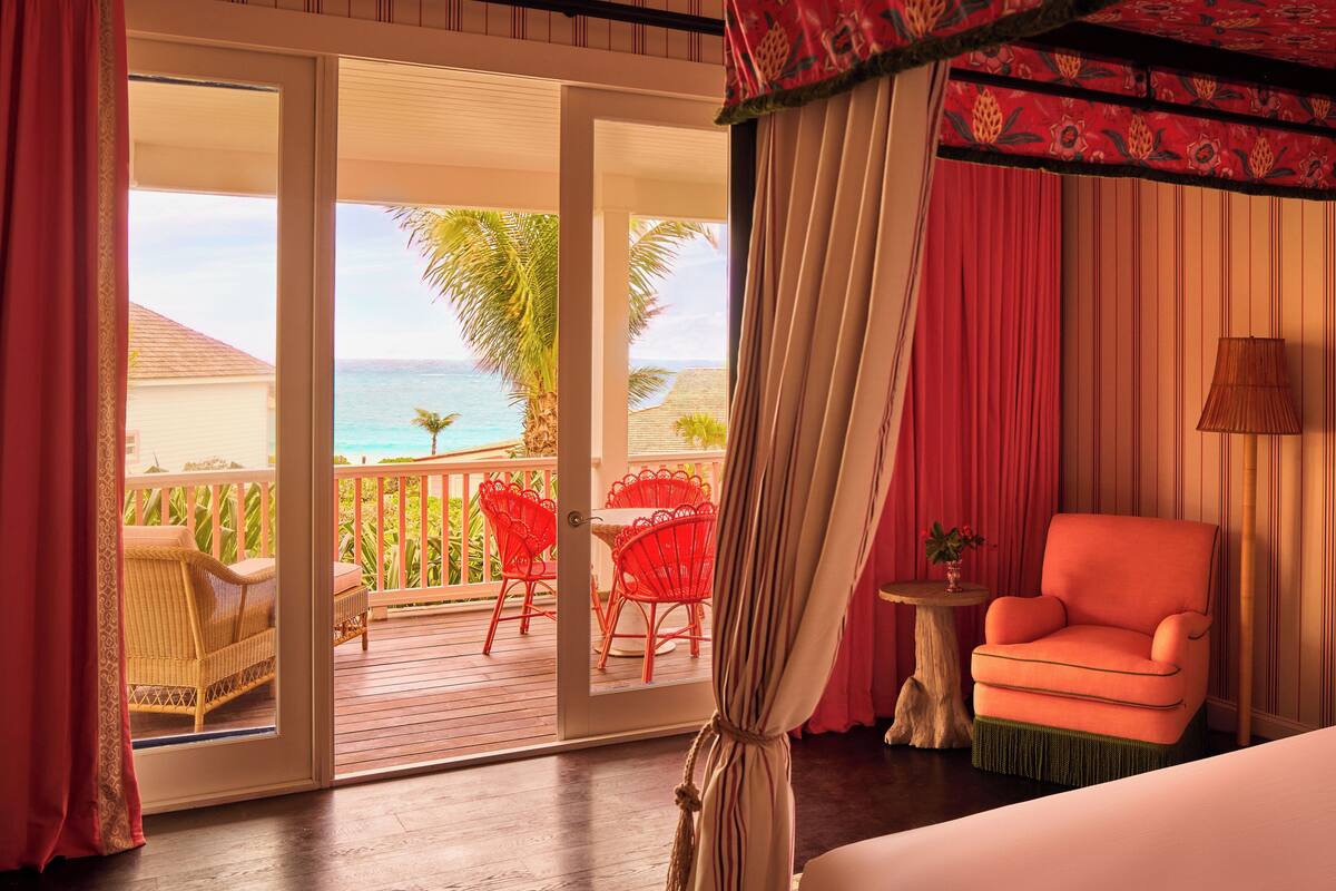 A room with a view of the ocean, a balcony with red chairs, and a pink armchair.