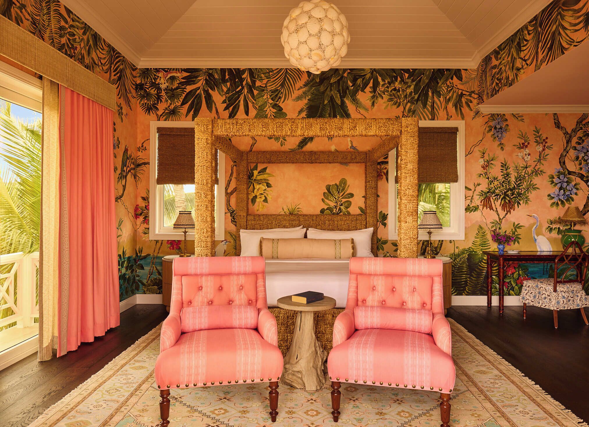 Une chambre à coucher sur le thème tropical, avec un lit, deux fauteuils roses, une table ronde en bois et une grande fenêtre avec des rideaux.