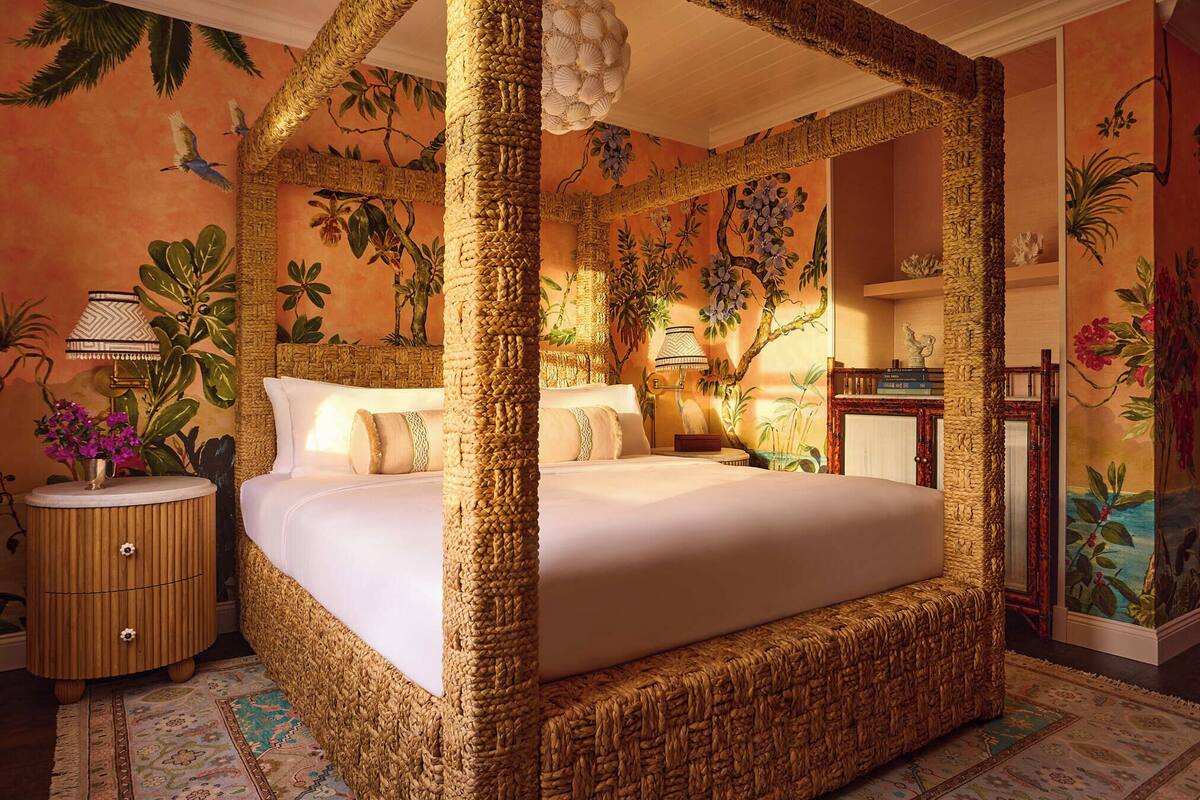 A four-poster bed with a woven canopy, a nightstand with a lamp, and a wall mural with tropical foliage.