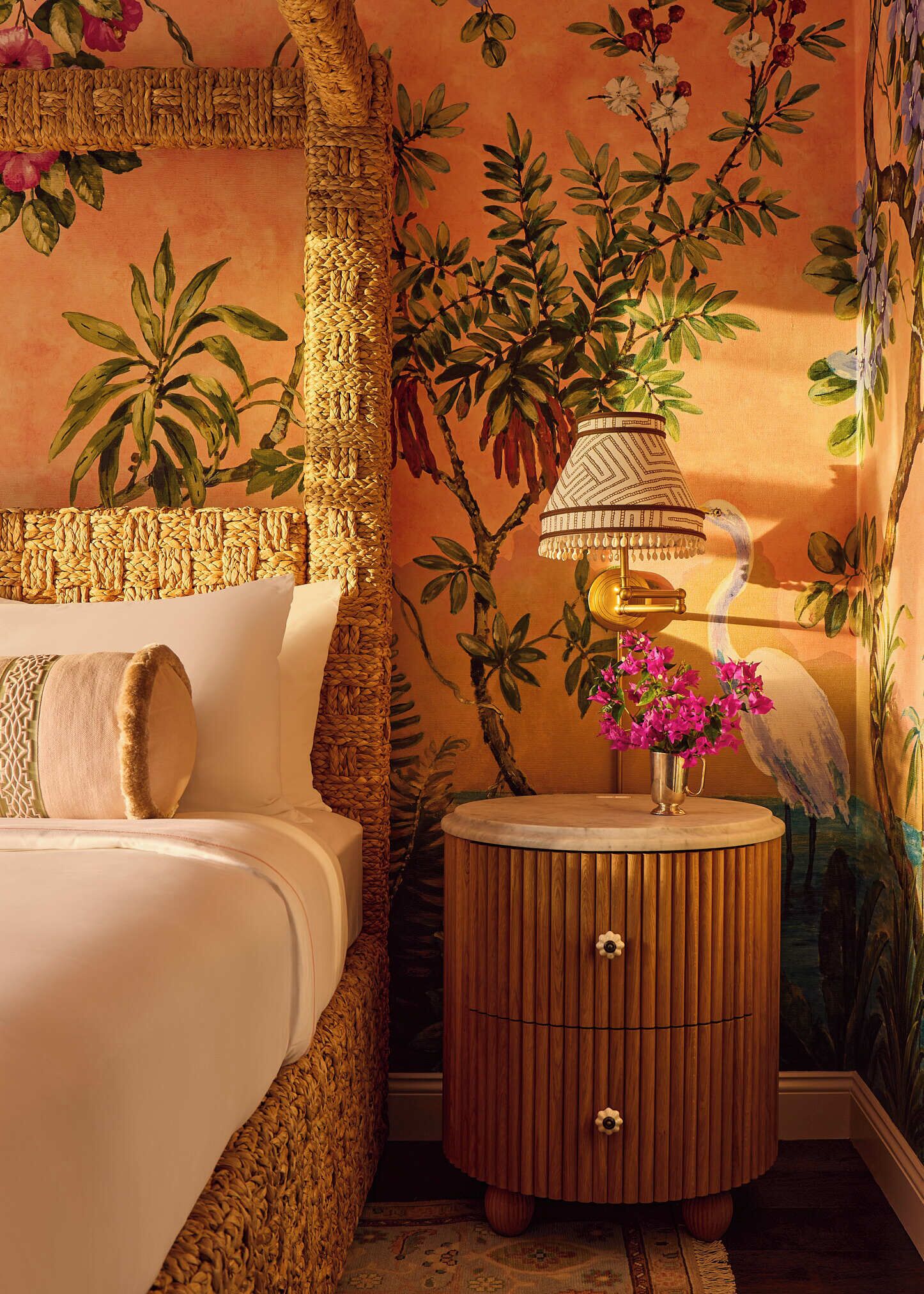 A bedroom with a bed, a round wooden nightstand, and a wall with a floral mural.