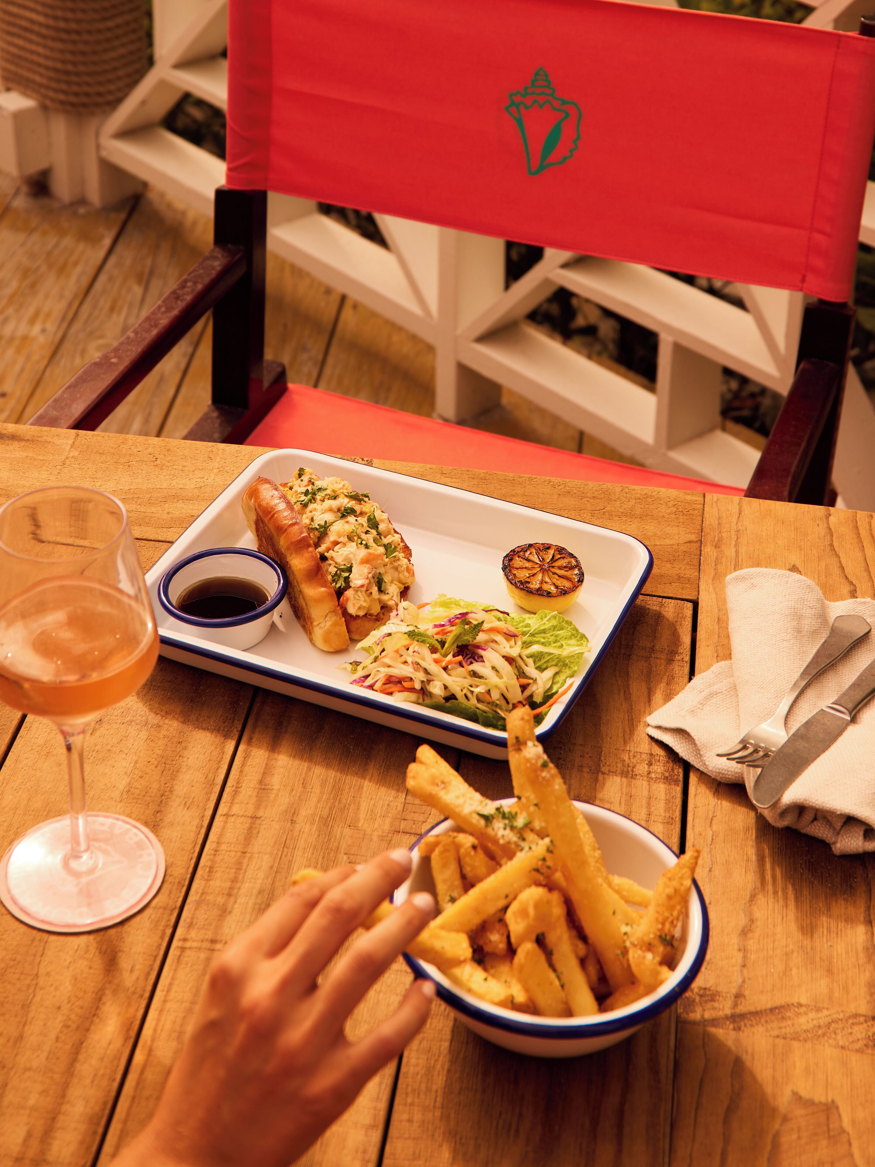 A wooden table with a white plate containing a sandwich, coleslaw, and a grilled vegetable. A glass of drink and a bowl of fries are also on the table.