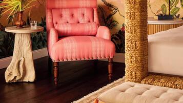 Una habitación con una butaca rosa tapizada, una mesa redonda de madera con un jarrón y un vaso, un pilar grande tejido y una cama con cabecera blanca.