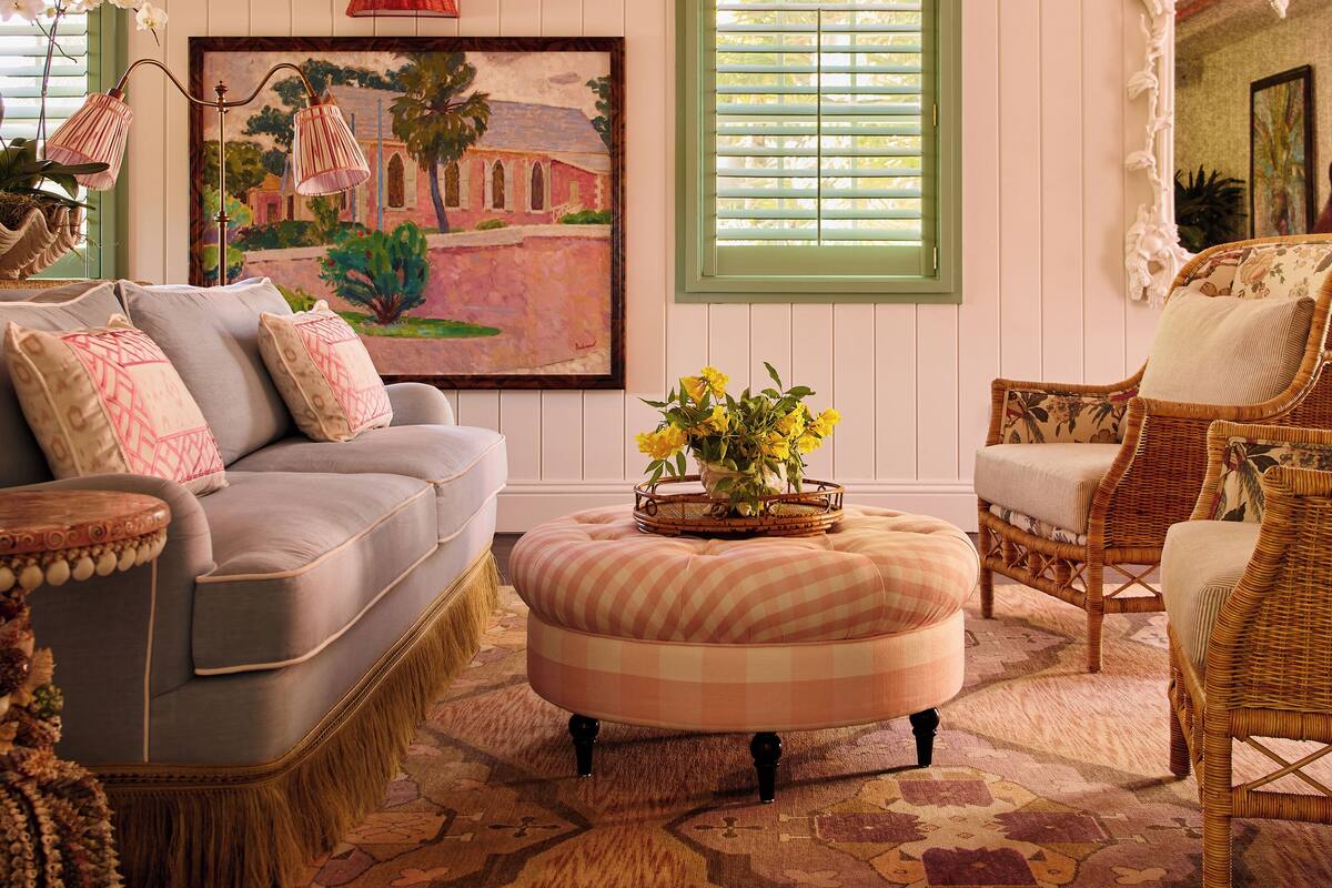 A cozy living room with a grey sofa, a round ottoman, wicker chairs, a ceiling fan, and a painting on the wall.
