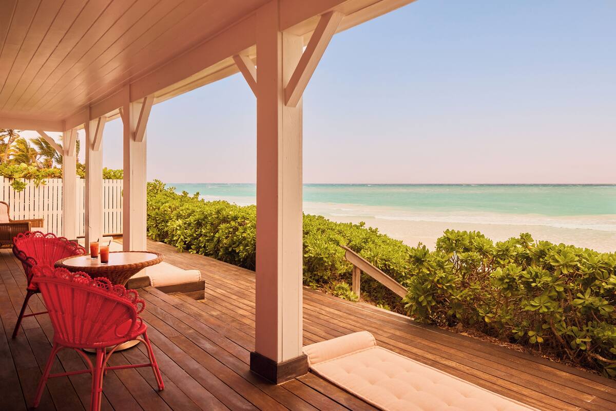 A wooden deck with red chairs and a table, overlooking a beach with clear turquoise water.
