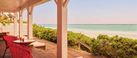 A wooden deck with red chairs and a table, overlooking a beach with clear turquoise water.