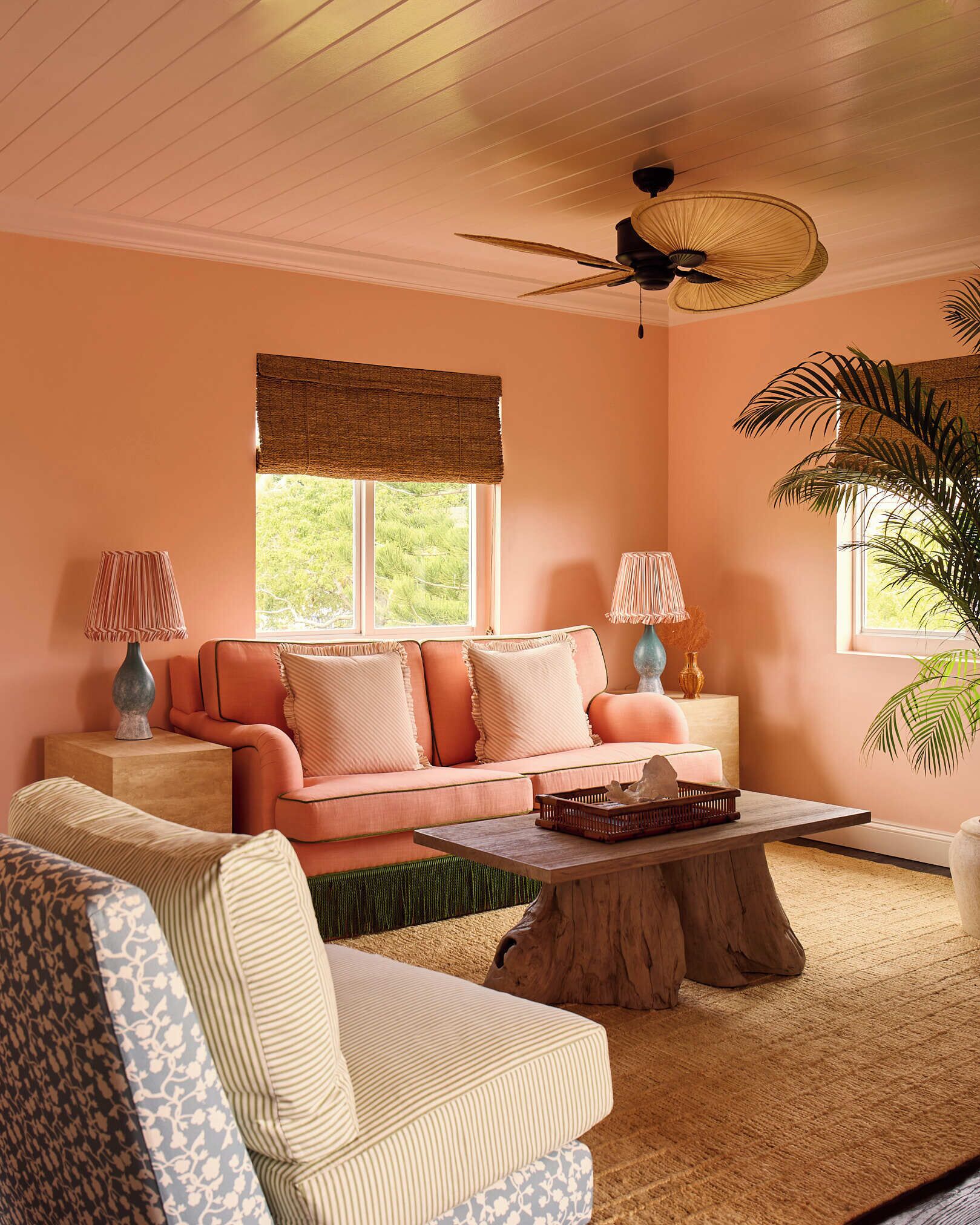 Un salon chaleureux avec un mur de couleur pêche, un ventilateur de plafond en bois, un canapé rose orné de coussins à volants, un fauteuil à rayures et une table basse en bois.
