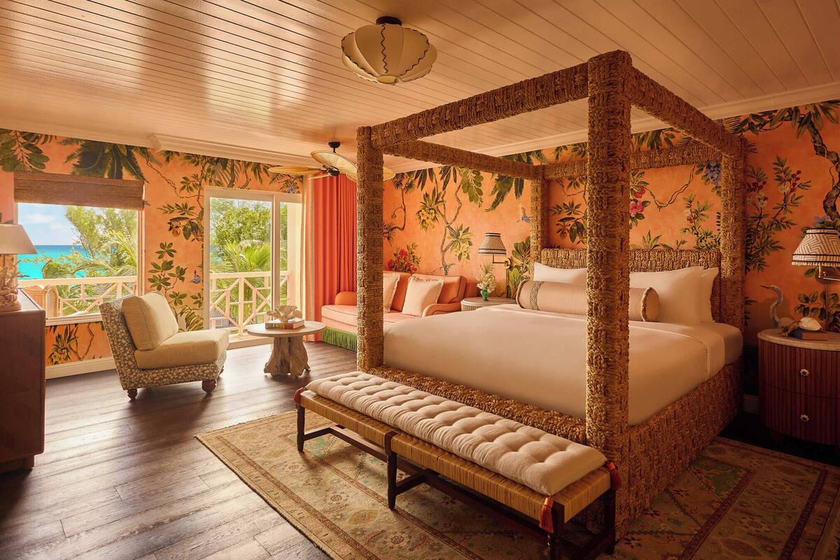 A four-poster bed with a woven canopy, a bench, a chaise lounge, and a view of the ocean through large windows.