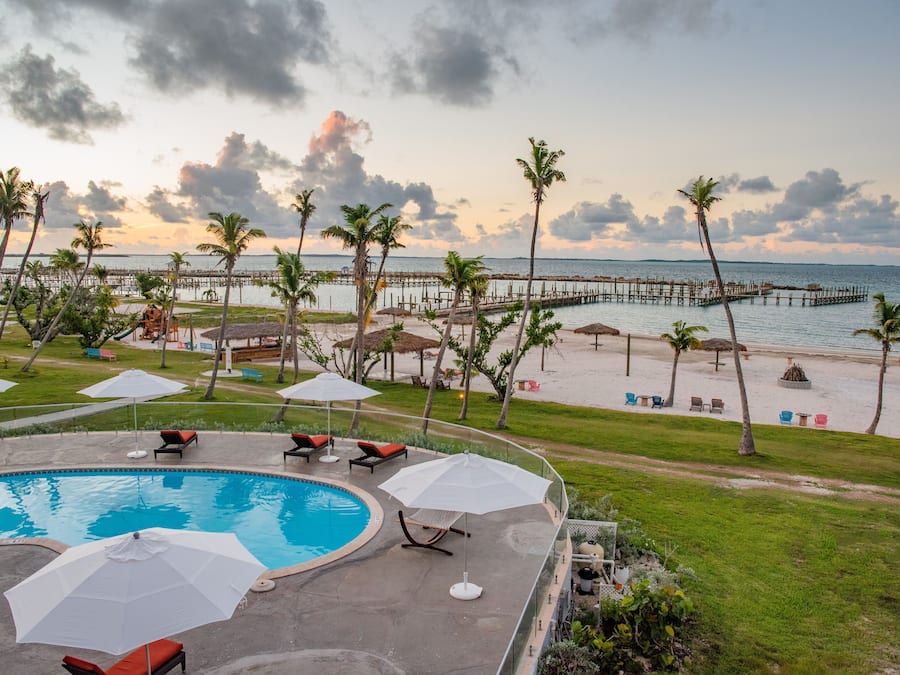 A pool area with lounge chairs and umbrellas, a beach with palm trees, and a pier extending into the water.