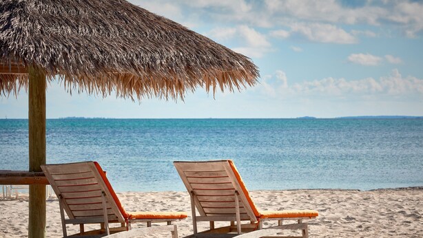 Deux fauteuils de plage en bois, dotés de coussins orange, sont placés sous un parasol en chaume sur une plage de sable, avec l’océan en arrière-plan.