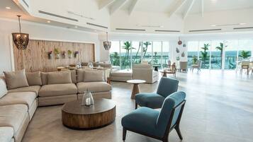 A spacious living area with a large sectional sofa, a round coffee table, and a blue armchair. The room features a wooden accent wall, a balcony with a view of the ocean, and large windows.