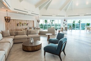 A spacious living area with a large sectional sofa, a round coffee table, and a blue armchair. The room features a wooden accent wall, a balcony with a view of the ocean, and large windows. - Abaco Beach Resort and Boat Harbour Marina (Marsh Harbour)