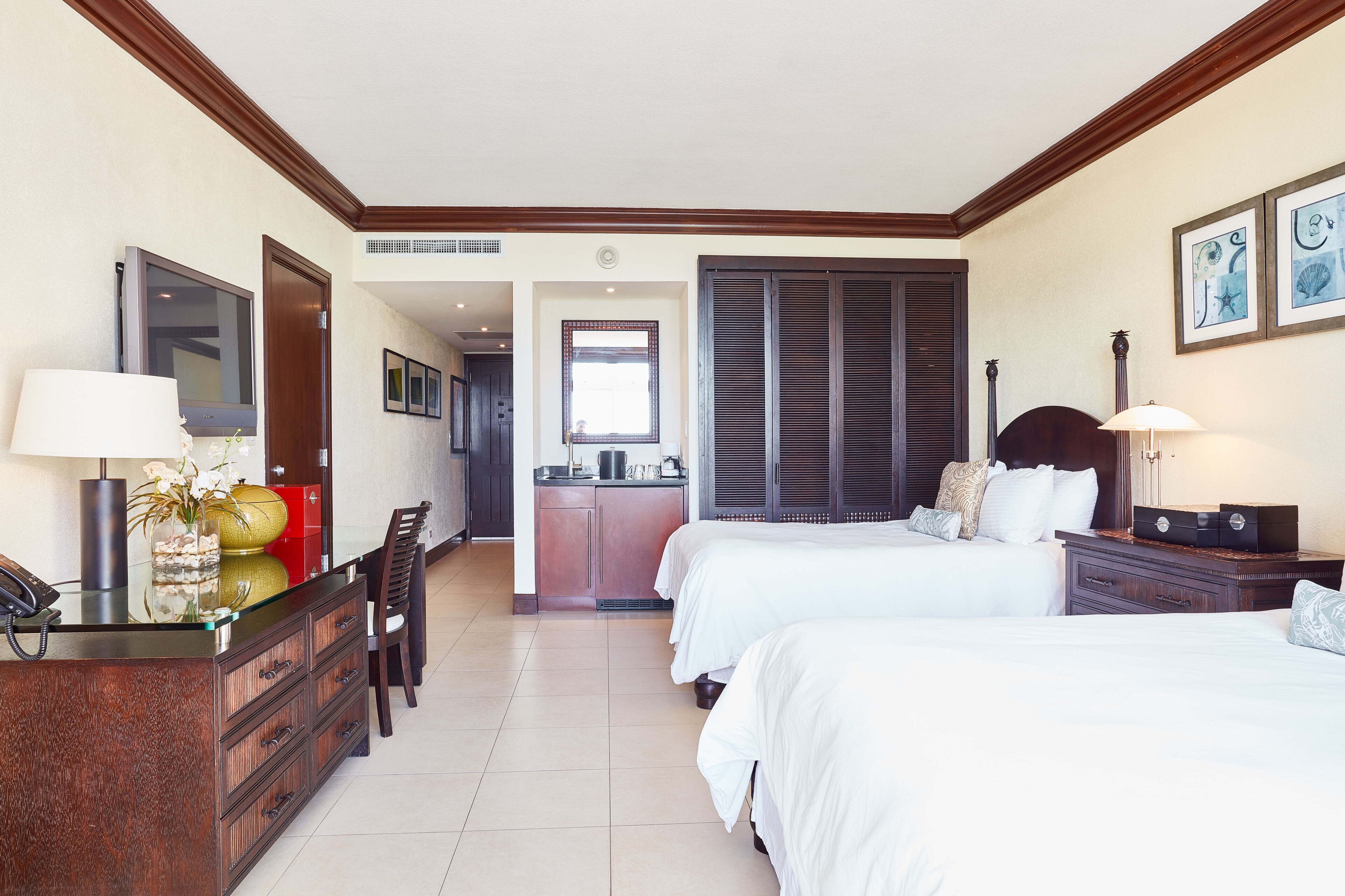 Habitación de hotel con dos camas, un escritorio con teléfono, una kitchenette pequeña y una ventana con persianas.