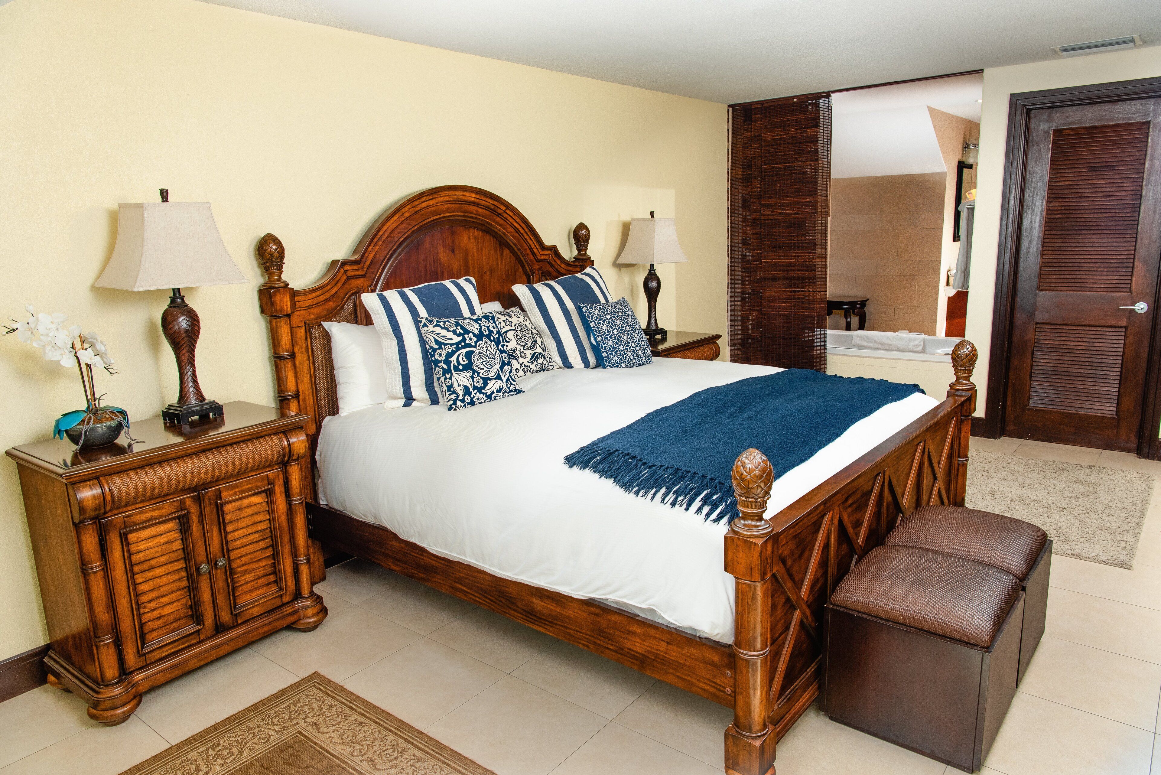 Une chambre à coucher bien rangée, avec un lit en bois, des tables de chevet, un banc et une salle de bain visible à travers une porte ouverte.