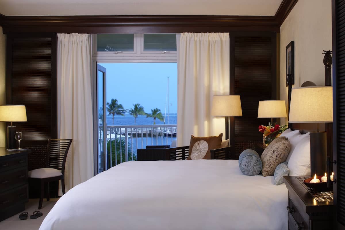 Habitación de hotel con una cama grande, dos mesitas de noche con lámparas, una silla, un aparador y vista al océano.